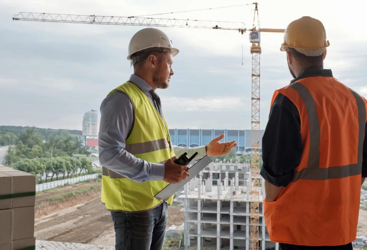 Técnico de prevención y encargado conversando a pie de obra frente a una grúa torre