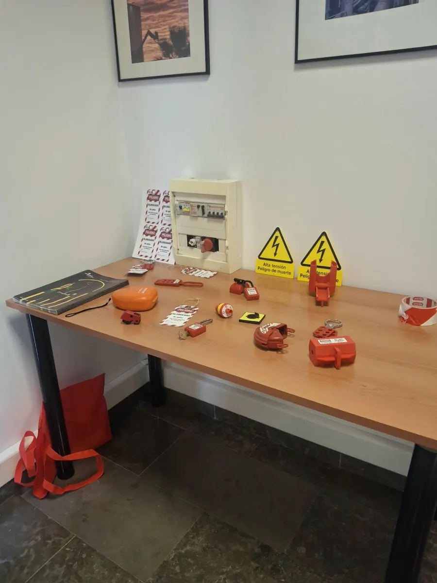 Mesa con material de bloqueo y etiquetado: candados, pinzas de bloqueo múltiple, tarjetas de peligro, un cuadro eléctrico de prácticas y señales de riesgo eléctrico